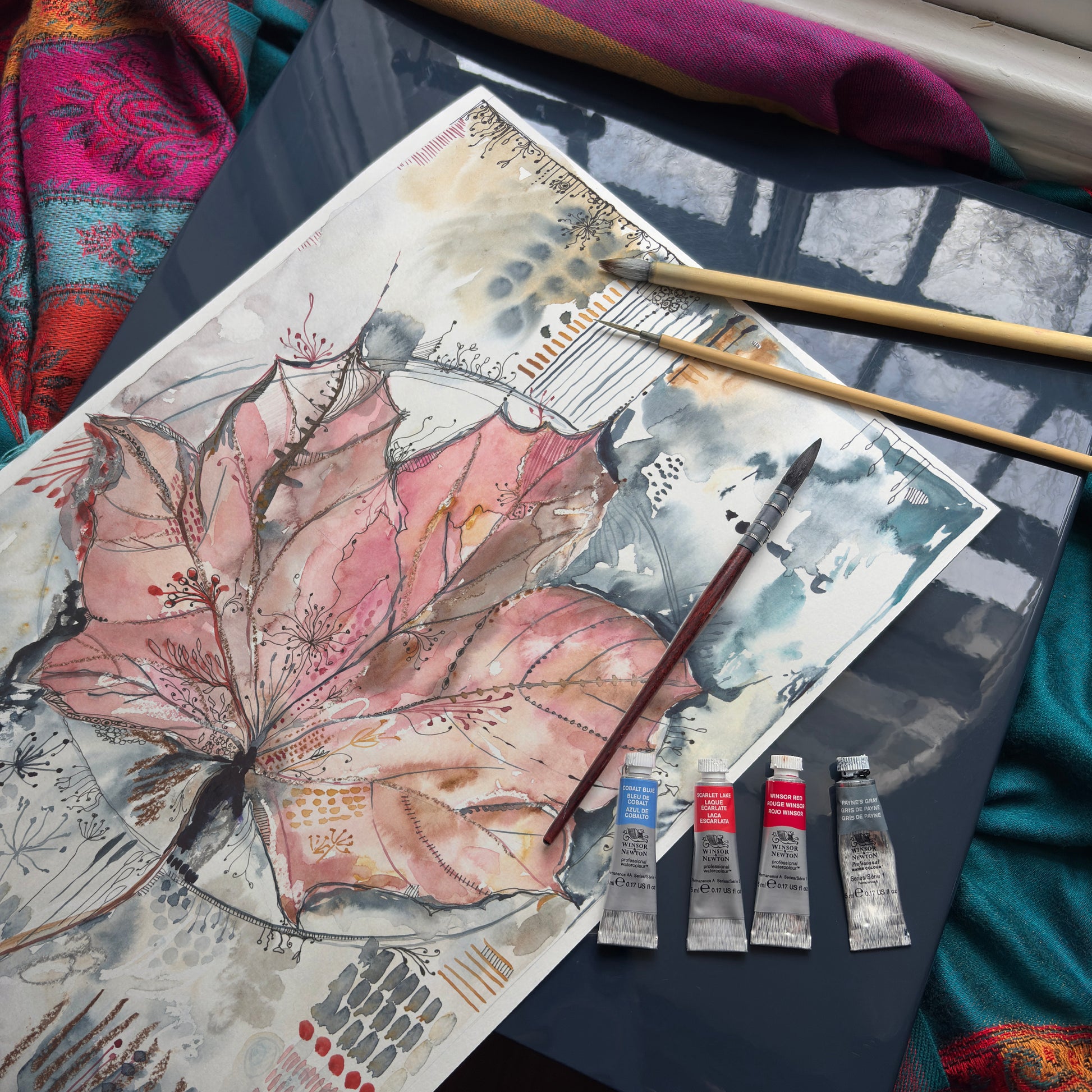 Finished painting of a maple leaf with painting supplies on desk in the sun.