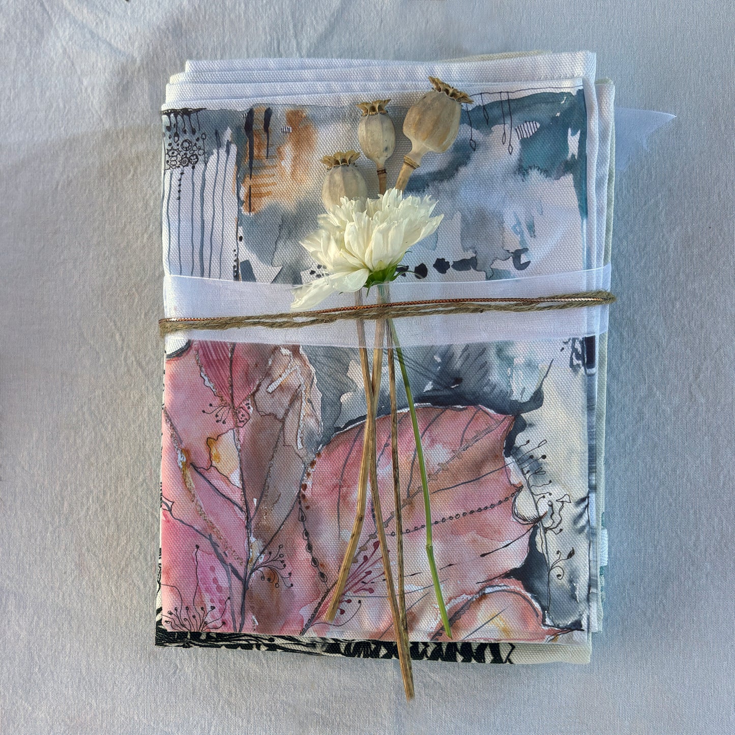 Folded tea towel, gift wrapped with ribbon, cosmos flower and dried poppy seed pods.