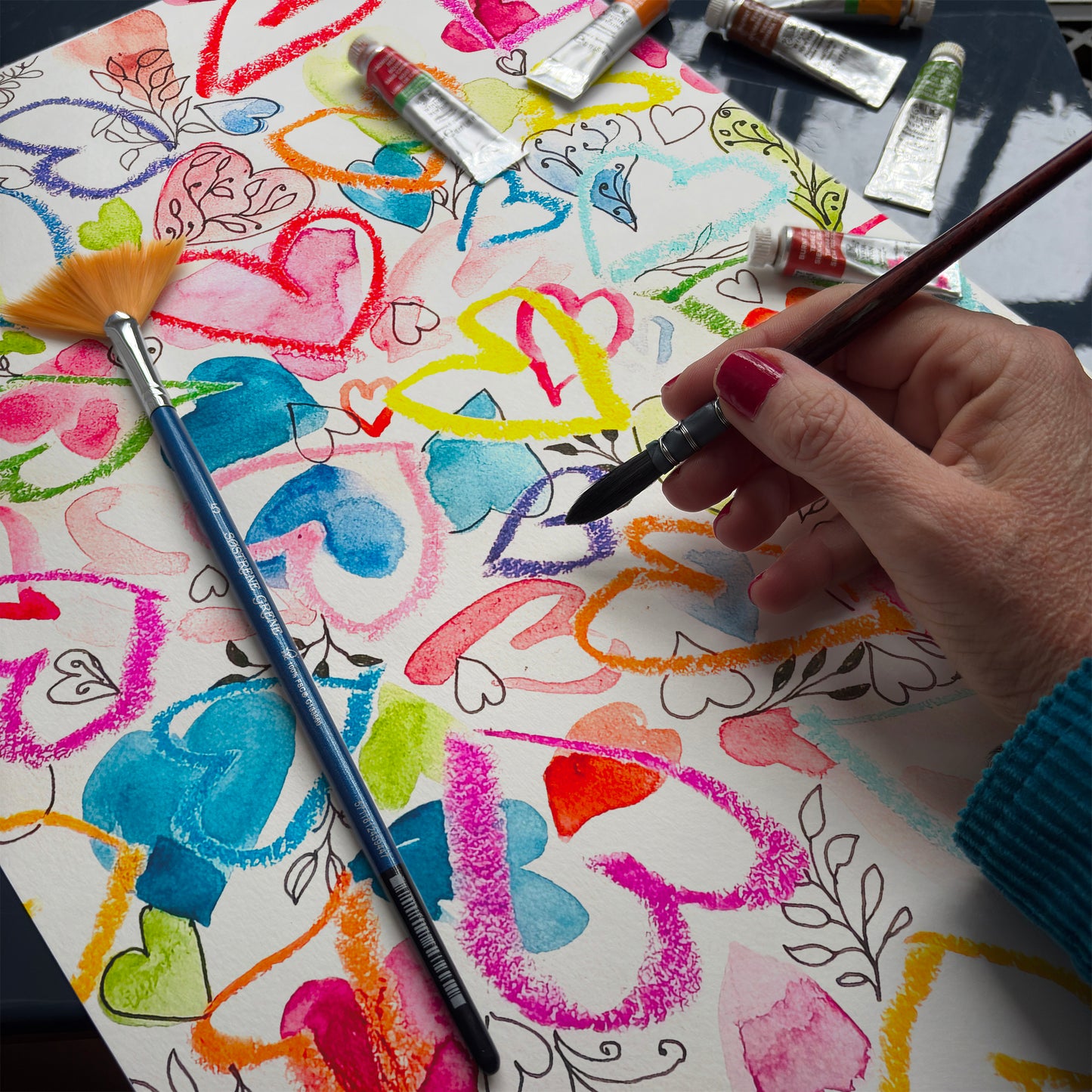 Rainbow painting with hearts on paper, surrounded by paint tubes and brushes.