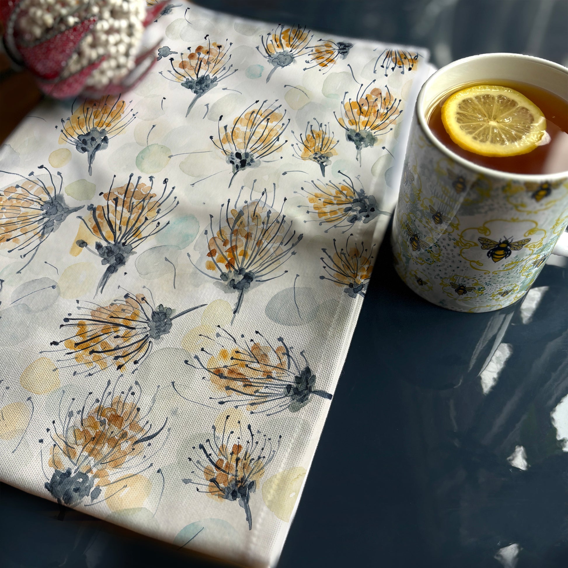 A tea towel with a watercolor-style botanical design featuring pincushion proteas in shades of gold, gray, and black, along with soft, abstract circular shapes in muted tones. The towel is laid out on a reflective dark surface. Beside it, there is a cup of tea with a slice of lemon, in a white mug decorated with a bee pattern. Sunlight casts soft shadows and highlights the texture of the fabric.








