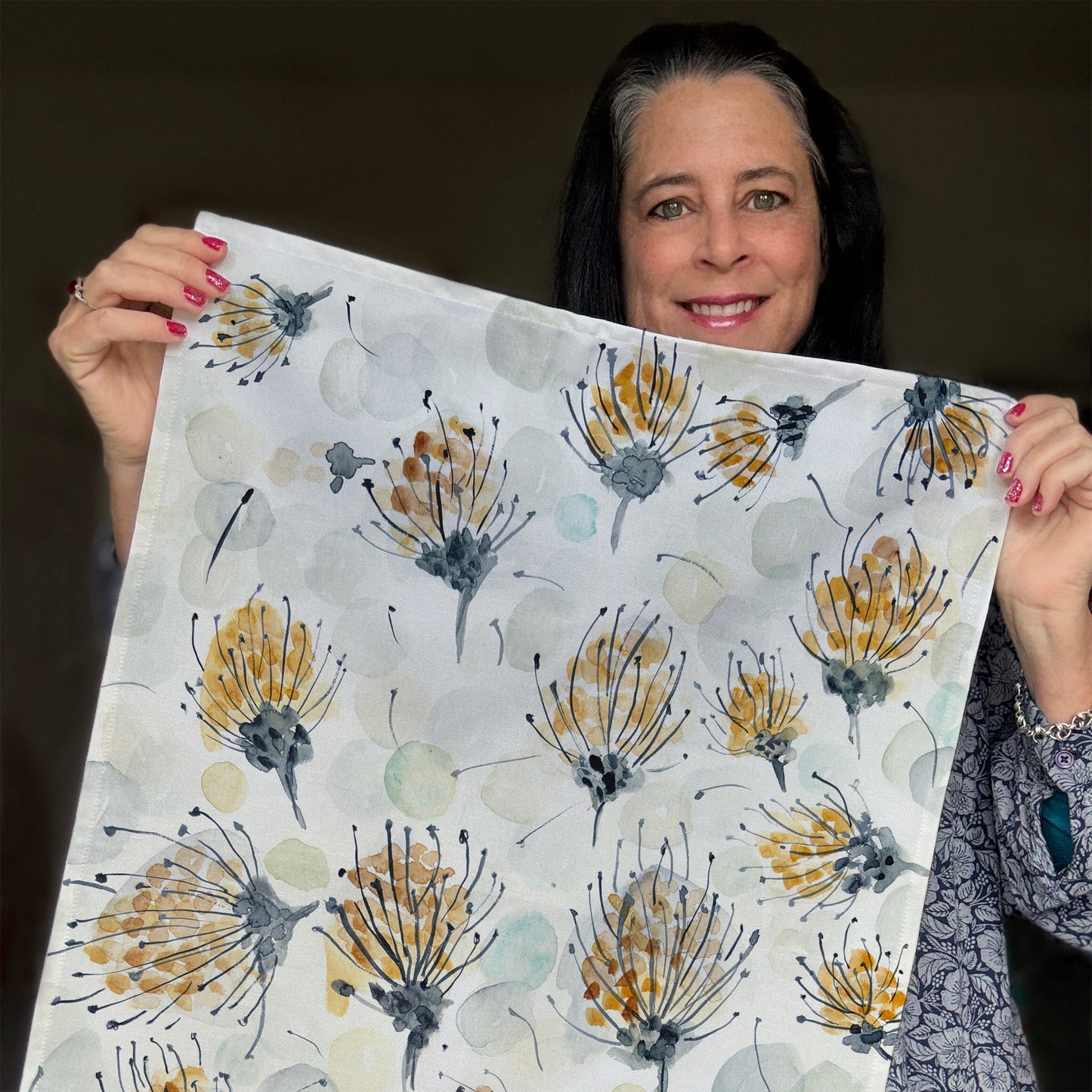A tea towel with a watercolor-style botanical design featuring delicate pin cushion protea in shades of gold, gray, and black, along with soft, abstract circular shapes in muted tones, held by Bonny.