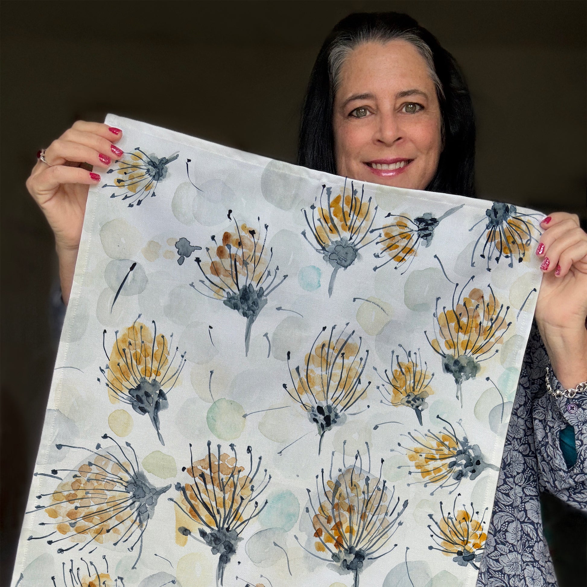 A tea towel with a watercolor-style botanical design featuring delicate pin cushion protea in shades of gold, gray, and black, along with soft, abstract circular shapes in muted tones, held by Bonny.