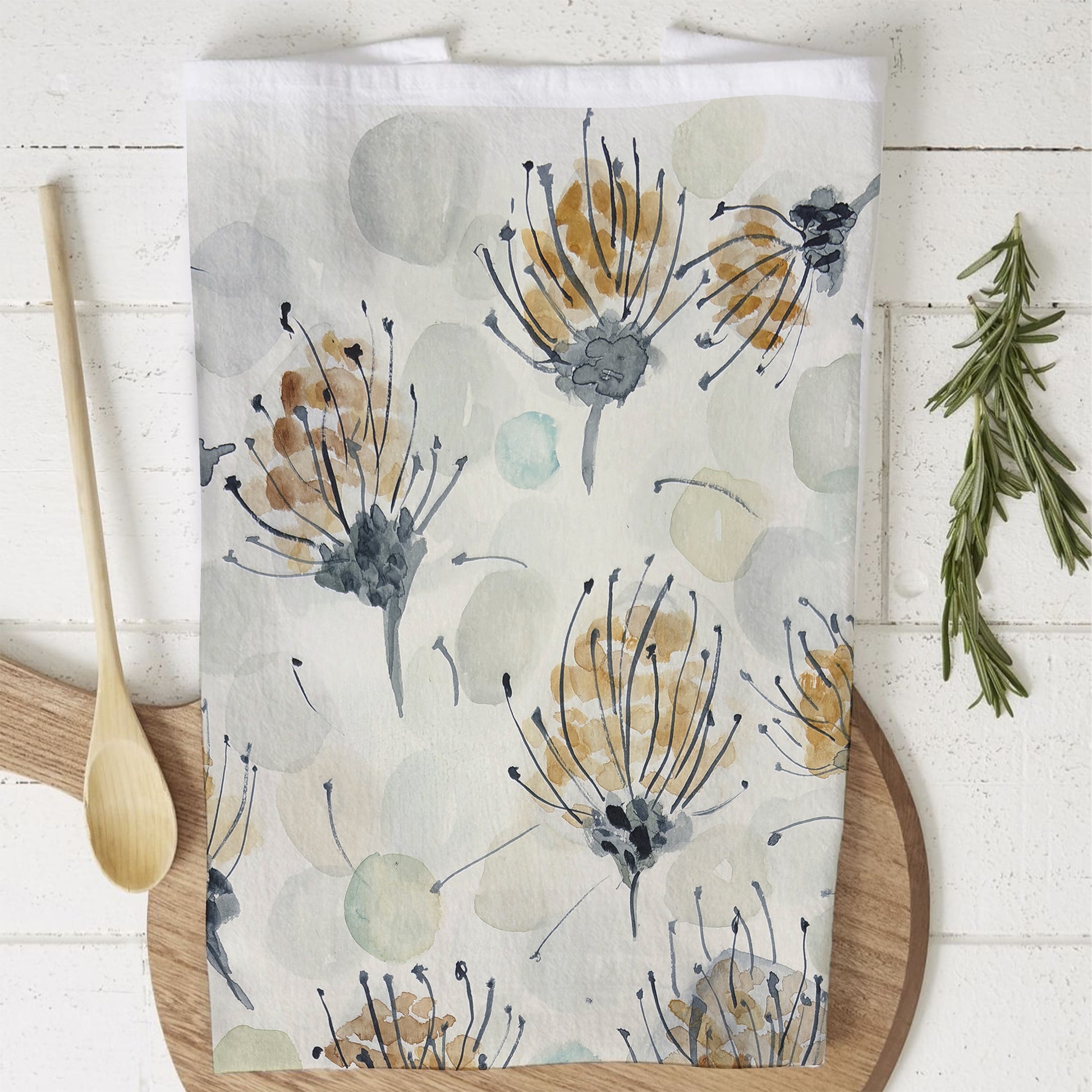 A tea towel with a watercolor-style botanical design featuring delicate pin cushion protea in shades of gold, gray, and black, along with soft, abstract circular shapes in muted tones. The towel is draped over a wooden cutting board on a white wooden surface. A wooden spoon and a sprig of fresh rosemary are also visible nearby.